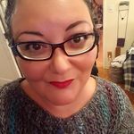 A close-up of a brown-skinned woman with red lipstick and perfectly arched eyebrows, looking into the camera with a playful gaze. They are wearing a grey sweater with stripes of blue and purple, black-rimmed glasses, and their dark hair pulled back from their face.