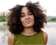 Portrait of jazmín calderón torres taken outdoors in front of a lake. jazmín, a trans-nonbinary Boricua, faces forwards and looks directly at the camera with dark brown shoulder length curly hair, a small patch of facial hair on their chin, septum and upper lip piercings. A tattoo on their sternum peeks through a green sleeveless shirt.