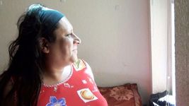 Still from the short film La Promesa (2016) by Adriana Kin. Adriana Kin is trans, Latina, and has long dark hair. The still shows Adriana's side profile as she looks to the right towards a window in a room with bare, pale walls.