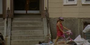 A child in front of a stoop on a bike. They are wearing a pink top, pink shorts, a light purple and black helmet, and a pink and light purple bike. There is a light blue bike on the ground behind them, as well as some cardboard boxes and trash bags on the sidewalk in front of them.