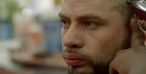 A close-up of someone getting their haircut, seen from the right side of their face and head. They have light skin and a thin beard, and are looking straight forward with an intense expression. At the top right you can see silver and red clippers trimming the hair on the sides of their head. The background is blurred out.