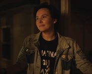 Still of June Thiele from an unidentified short film. June has light skin, short dark coiffed hair, a small silver nose ring, and is wearing a denim jacket over a black shirt. They are standing in front of a non-descript building at night, their face bathed in warm light as they stare wistfully at something past the camera.