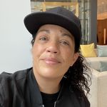 Raven Davis is a mixed race Indigenous 2-spirit person with brown eyes, light skin, freckles and dark curly hair tucked under a ballcap. A couch and high-end interior are visible behind them.