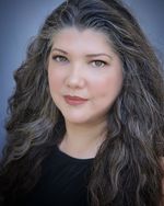 Studio portrait of Alexandra Sandy Harjo-Livingston who is of Chickasaw, Tsalagi, and Mvskoke descent. He has long, dark wavy hair with a few streaks of grey, light-skin, and brown eyes. They are wearing a black t-shirt and a neutral shade of lipstick.
