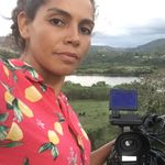 Arisleya Dilone stands beside a video camera in front of a lush landscape. Arisleya is a Latinx/hispanic person with brown skin and wavy or curly hair pulled back. She wears a vibrant button-up with a lemon print.