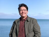 Finn Simard is an Indigenous person with clear glasses, short black hair, light-skin, and a slight mustache. He wears a light jacket over a button-up shirt. He stands in front of a body of water.
