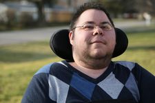 Dom a person with green eyes and brown spiked short hair gives a small smile. They are wearing gold wire rim glasses, and have some stubble and hair along his cheek line. This headshot shows from his mid chest area upward. He is wearing a blue, black, and gray Argyle long-sleeve shirt. He sits in his wheelchair, and his black headrest wraps around the back of his head. The background is blurred, but you can tell there is grass behind him and he is outside.
