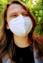 Sandra Alland has brown hair past their shoulders and light-skin. They wear an N-95-style mask, black t-shirt and blazer. They are pictured outdoors with leaves visible behind them.