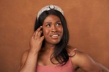 Ava Davis is trans and black with brown skin featured in a shiny stone covered crown and a spaghetti strap tank top. She is looking up and to the right of the image smiling and has her left hand to her face touching her hair. She has dark brown chest length straight hair in front of a brown background.