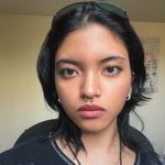 Ella Saini is Filipino-Indian with light skin. They have neck length black hair with a pair of sunglasses pushed back sitting on her head. Ella has a septum piercing and two earrings. They are shown from just under the shoulders up wearing a black shirt with white bleach marks on it with a necklace that is out of frame. Ella stares intently and a little bit aggressively into the camera.

