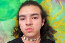 Jamie John with a neutral expression in front of a mural. Jamie is an Anishinaabe and Korean-American person with light skin, some facial stubble, and curly hair to his shoulders. They wear a black t-shirt and choker of large beads.