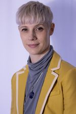 Lora Campbell with a slight smile. Lora has light skin, a platinum bowl cut, and subtle makeup. They wear a pendant necklace, a turtleneck and yellow blazer.
