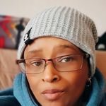 Lynnell Stephani Long is Black and Intersex with light brown skin. She is shown from her chin up in a white beanie and a blue hoodie with small rectangular glasses. They are on a couch wearing small hoop earrings with a white wall behind the couch.