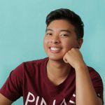 Marco Chiong is a Trans Asian-American with light tan skin in front of a light blue background. They are wearing a maroon short sleeve t-shirt that has white lettering on it. Marco is resting their face on their right fist smiling. They have black hair with the sides faded and short hair on the top.