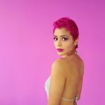 María José Maldonado looks over her shoulder into the camera. María is a Salvadoran-Ecuadorian person with short pink hair and brown skin. They wear a halter top and fuchsia lipstick and eye shadow. She stands in front of a bright background of pure fuchsia.