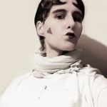 Black and white photo of Marlowe Mitchell referencing Claude Cahun. Marlowe has short hair styled with heart bangs, a pale face with hearts on his cheeks and lipstick applied in a heart. They wear a white shirt and scarf.
