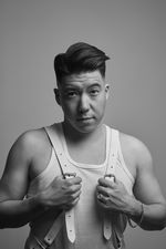 Black and white photo of Max Disgrace with raised eyebrows. Max is of Chinese descent with a precise undercut. He wears a tank top, leather suspender, heavy chain bracelet and larger ring on his wedding finger.