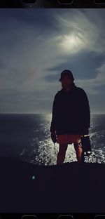 Silhoutte picture of MG Evangelista in front of a body of water, a sun, and wispy clouds. Most details of MG are left in darkness but we can make out that they have short hair under a hat, are wearing long sleeves and shorts, and hold a bulky light.