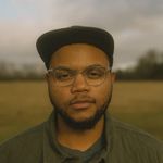 Remy Styrk is a Black trans man with light brown skin in front of a field that's blurred out. He is wearing a dark green hat with clear glasses. He has a beard and is wearing a dark green jacket with a gray shirt underneath.