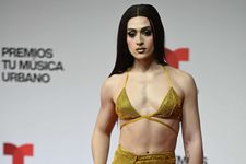 Villano Antillano is a transfemme Puerto Rican woman. She has light skin in front of a white background. She is wearing a yellow bikini top with yellow shiny jewels on top of it and matching pants. She has long straight black hair going down her back. She has her makeup done with blush on her cheek bones and black eyeliner.