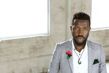 Pages Matam is a Black person and genderqueer with dark brown skin posing in front of a concrete wall with windows. They are wearing a gray suit with a pin of Africa with the Black Liberation Flag colors on it. They are wearing a white undershirt and necklace that looks like a hook. They have short curly black hair, a beard and a green-black pocket square.