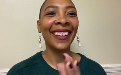 Jasmin Benward is Black with light brown skin. She is smiling, wearing lipstick, has a buzzed head and wears dangly earrings. They are posing in front of a white wall in a dark green blue sweater. 
