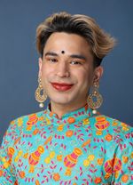Shiva Raichandani is nonbinary South Asian with brown skin. They are posed in front of a dark blue background wearing a light blue shirt with orange and pink designs covering it. They have short curly ombre hair with gold earrings, a nose piercing, and a piece of jewelry on their forehead. Their wide, closed-lipped smile is accentuated by their bright red lipstick.