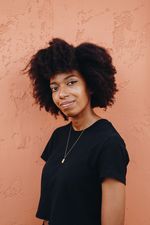 Olivia Peace is Black queer person with brown skin. She is posing in front of a clay wall with curly black hair with a nose piercing and a gold necklace. She is wearing a black crop top.