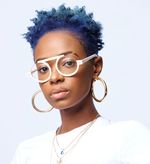 Morgan B. Powell is Black and non-binary with dark brown skin. They are posing in front of a white background in a white t-shirt and three gold necklaces. They have  white glasses and golden hoops and a nose piercing. They have short curly blue hair with the sides faded.