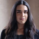 Ryan Rox is trans and non-binary with light skin. They have long straight brown hair and are posing in front of a tan background in a black blouse.