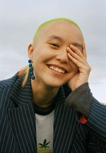 April Lin is gender fluid with light skin and is posed in front of a light gray background. They have a lime green shaved head wearing a navy blue striped blazer over a white graphic t-shirt.
