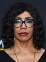 Kase Peña is a Latinx trans woman with light brown skin. She is posing in front of a black background with curly black hair at shoulder length. She has black glasses, gold hoops, a gold necklace and a black t-shirt.