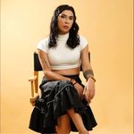 Rod Singh is trans femme with light brown skin. She is sitting in a directors chair in front of a pastel yellow background. She is wearing a white crop top and a long black skirt with her legs crossed. She has shoulder length curly black hair.