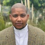 Sydney Baloue is a transgender South Asian Black man posed in front of some bushes. He is bald and wearing a white button up and a blazer over it.