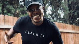 Sir Lex Kennedy is a Black trans masculine person with dark brown skin posed in front of a brown fence with trees poking over it. He is wearing a black baseball hat and clear framed glasses. He has a septum piercing and is wearing a black short sleeved t-shirt that says, Black AF.