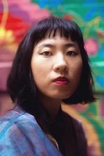 Jess X Snow is nonbinary and Chinese with light skin posed in front of a blurred out graffitied wall. They have black hair in a bob with the back longer than the rest and bangs. They have a septum piercing and they are wearing a colorful jacket.