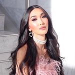 Julie Vu is an Asian-Canadian trans woman with light skin posed in front of a gray staircase. She has long curly dark brown hair with earrings in a heart shape made of pearls. She is wearing a choker made of pearls and a pink feather top.