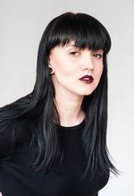 A soft, classic studio portrait of Dayna Danger against a professional light gray backdrop. Dayna Danger is fair skinned Métis, Saulteaux, and Polish person. They have bold black mid length hair with full bangs, black spacer earrings, light face makeup with a statement gold inner eye corner, bold red lip, a gold nose ring and silver monroe piercing. They are wearing a simple black long sleeve shirt.