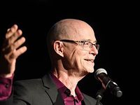 John Killacky in his 70s grinning and giving a speech on a stage. John is a white cis gay man with a bald head and slim rectangular glasses. He is wearing a black suit with a plum coloured dress shirt.