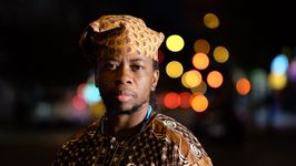 Brown skinned Yoruba person wearing brown and gold traditional nigerian attire, with Gold and Burundy hat over braided locs, and short beard looking forward at the camera. Queer, Yoruba Black person using they/them pronouns.