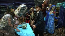 A bunch of people stand around in various costumes. The person in the front has light skin, a single blue goggle on their head, and is wearing an opaque black polka-dot rain poncho. The person's hands rest on a half-circle table with a blue fabric cover and a fishbowl resting on it. The person on the other side of the table wears a navy-blue suit and a black top hat, and has a white bushy mustache and eyebrows, and light skin. The other costumed people in the background appear to be in motion.
