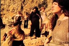 A bunch of people standing in the desert. The person closest to the camera has long straight brown hair, white skin, is wearing a chain vest, has a bandana on their forehead, and is looking up at the sun. The background people are scattered, with some looking up at the sun and others looking off in other directions.