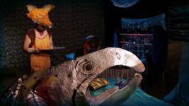 A silver creature with an open, beak-like mouth and yellow eyes appears in profile at the forefront of the image. In the background, someone in an orange fox costume holds a serving platter. The silver creature is leaning on a table which is covered in a blue table cloth and has a golden clutch purse sitting on it.