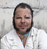 Harvey Rabbit is seen from the shoulders up, in front of a concrete wall with peeling white paint. He has short, curly black hair and light tan skin, with prominent mutton chops on the sides of his face, a large mustache above his lips, a small soul patch below his lips, and a clean-shaven chin. His lips are painted in a vibrant dark pink which accentuates his slight smile. On his neck, he wears a heart-shaped choker which is pink on the left and light blue on the right, evoking the trans flag. A small blue crystal dangles from the center of the choker. His shirt is white and ruffled, like that of a buccaneer.