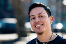 Anto(n) is a Chilean-American person with short, straight brown hair, brown freckled skin, and a light moustache and goatee. They grin at the camera in a friendly way. They are wearing a metal chain necklace and a black T-shirt. Behind them is a blurred out urban scene.