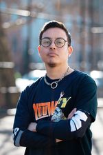 Anto(n) is a Chilean-American person with short, straight brown hair, brown freckled skin, and a light moustache and goatee. They are wearing round black glasses and they look at the camera in a challenging way. They wear a metal chain necklace and a black t-shirt over a black long-sleeved shirt with colorful writing on it. Their arms are crossed. Behind them is a blurred out urban scene.