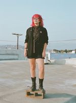 Full body creative photograph of Sam Gurry, a white nonbinary queer person with a neon pink shaggy mullet and matching bold sunglasses. They are wearing a dark blazer, a striped button-up shirt, dark shorts and boots, and are posed standing on top of a concrete cinder block on the roof of a lowrise building in Los Angeles.