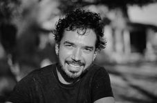 A black-and-white photo of Ryan Trecartin, a white person with dark curly hair and beard, smiling warmly at the camera. The background is slightly blurred, giving the image a soft feel.