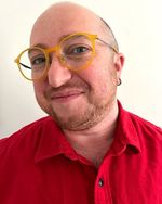 Selfie taken by Ezra Berkley Nepon against a plain white wall. They have light skin, a red beard, and short, closely-shaved hair. They are wearing round, bright yellow glasses and a red collared shirt accessorized with small hoop earrings.