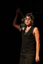 Adelaide Windsome stands on black theatre stage looking directly at the camera. She has short wavy hair and is wearing an all black outfit of a vest, dress pants and glasses. Her right hand is raised above her, holding a grey-and-white plush figure that rests on her shoulder. She is illuminated by a stage spotlight which leaves the background in the darkness.